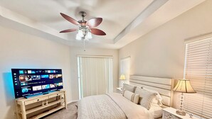 A bedroom with a bed, a TV on a wooden stand, a ceiling fan, and a bedside table with a lamp. - Grand Village 4BR Pool Home near Disney World (Davenport)
