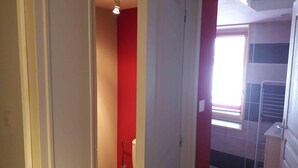 Een smalle gang die leidt naar een badkamer met een rode accentmuur, een toilet en een raam met zonwering.