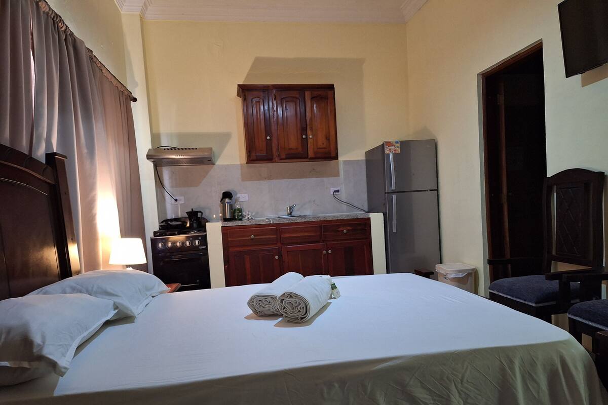 Ein Hotelzimmer mit Bett, Schreibtisch, Stuhl und einer Küchenzeile, die einen Herd, einen Kühlschrank und Holzschränke beinhaltet.