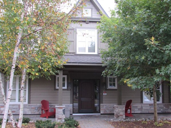 A two-story house with a dark brown front door, a small porch with two red Adirondack chairs, and a stone foundation.