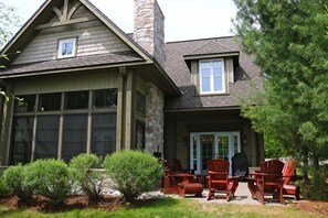 A two-story house with a stone chimney, a garage with large windows, and a patio with wooden chairs and a table.