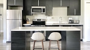 Une cuisine moderne avec des meubles de couleur sombre, des appareils en acier inoxydable, un îlot central doté de deux tabourets de bar et un micro-ondes encastré.