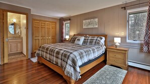 A bedroom with a wooden bed, a nightstand with a lamp, a window with a view of trees, and a door leading to a bathroom. - Heart of Killington location. Pet-friendly! (Killington)
