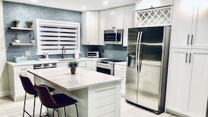 A modern kitchen with white cabinets, a stainless steel refrigerator, a built-in oven, and a microwave. There are two bar stools and a potted plant on the island. - Newly Furnished! Minutes from beach, heated private pool, Elegant and Coastal!   (Naples)