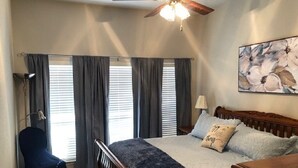 Ein Schlafzimmer mit Bett, Sessel, Schreibtisch, Deckenventilator und einem Fenster mit Vorhängen.