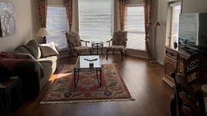 Ein Wohnzimmer mit einer Couch, Sesseln, einem Couchtisch und einem Fernseher. Zwei Fenster mit Jalousien und ein großes, rundbogenförmiges Fenster über der Couch.