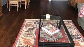 Ein Wohnzimmer mit einem Holzesstisch, Stühlen, einem Couchtisch mit einem Buch, einem gemusterten Teppich und einer Küche im Hintergrund.