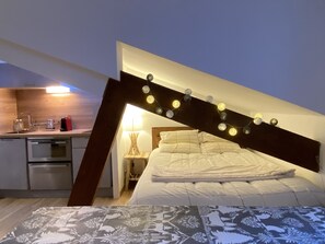 A compact bedroom with a sloped ceiling, a bed with white bedding, a bedside table with a lamp, and a kitchenette area with a stove and sink.