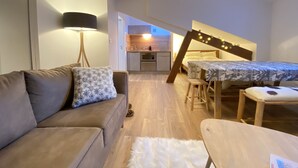 Un espacio habitable compacto con sofá, una mesita, una cama con escalera y una pequeña cocina en el fondo.