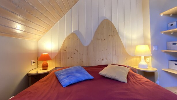 A bedroom with a wooden headboard, a red bedspread, two pillows, and bedside lamps.