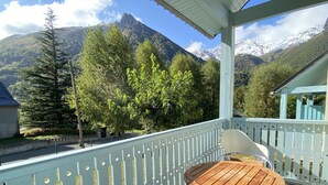 A balcony with a wooden table and chairs, overlooking a mountainous landscape with snow-capped peaks and lush greenery.