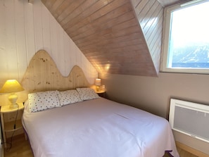 Quarto com teto inclinado, cama com almofadas de bolinhas, abajures e janela com vista para uma montanha.