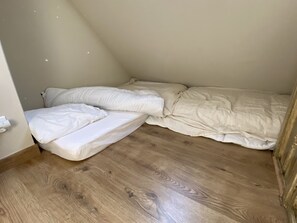 A small, single-occupancy room with a sloped ceiling, wooden flooring, and white bedding.