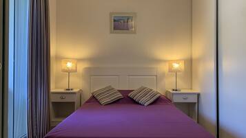 Una cama bien hecha con una colcha morada y dos almohadas, flanqueada por mesitas de noche con lámparas. Encima de la cama hay un cuadro enmarcado, y la habitación tiene un diseño sencillo y limpio con un ventanal grande a un lado.