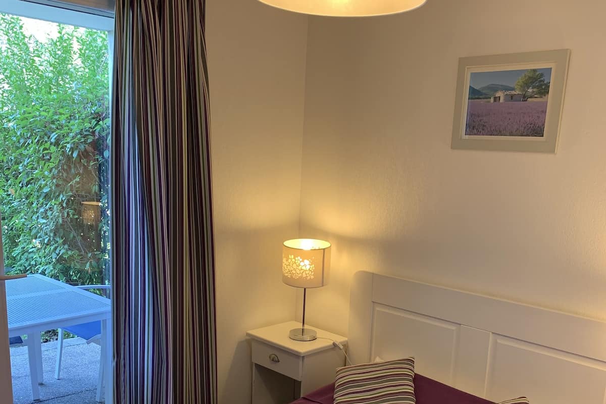 Une chambre à coucher avec une couvre-lit violette, des coussins à rayures, une lampe de chevet et un tableau encadré accroché au mur.