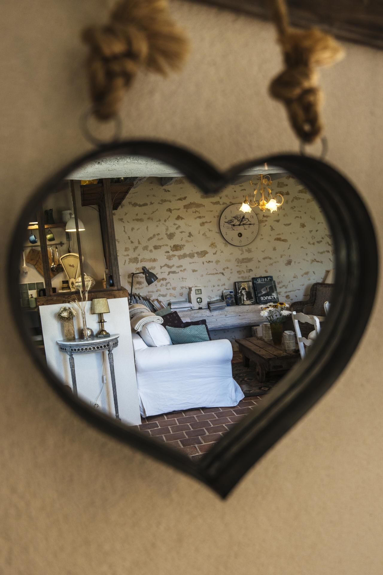 A heart-shaped mirror reflecting a living room with a white sofa, a small table, and a lamp.