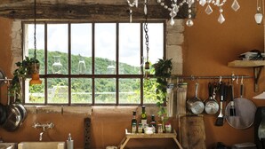 A rustic kitchen with a large window, wooden shelves, hanging pots, and a variety of kitchen utensils. - Le Domaine des Carriers - Gîtes (Chevroches)