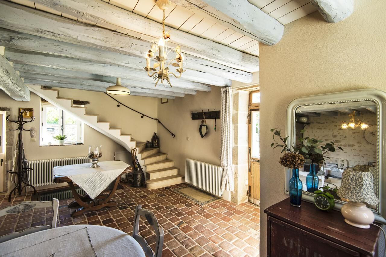 Um interior rústico com vigas de madeira aparente, uma escadaria, uma área de refeições com mesa e cadeiras, e um espelho com objetos decorativos.