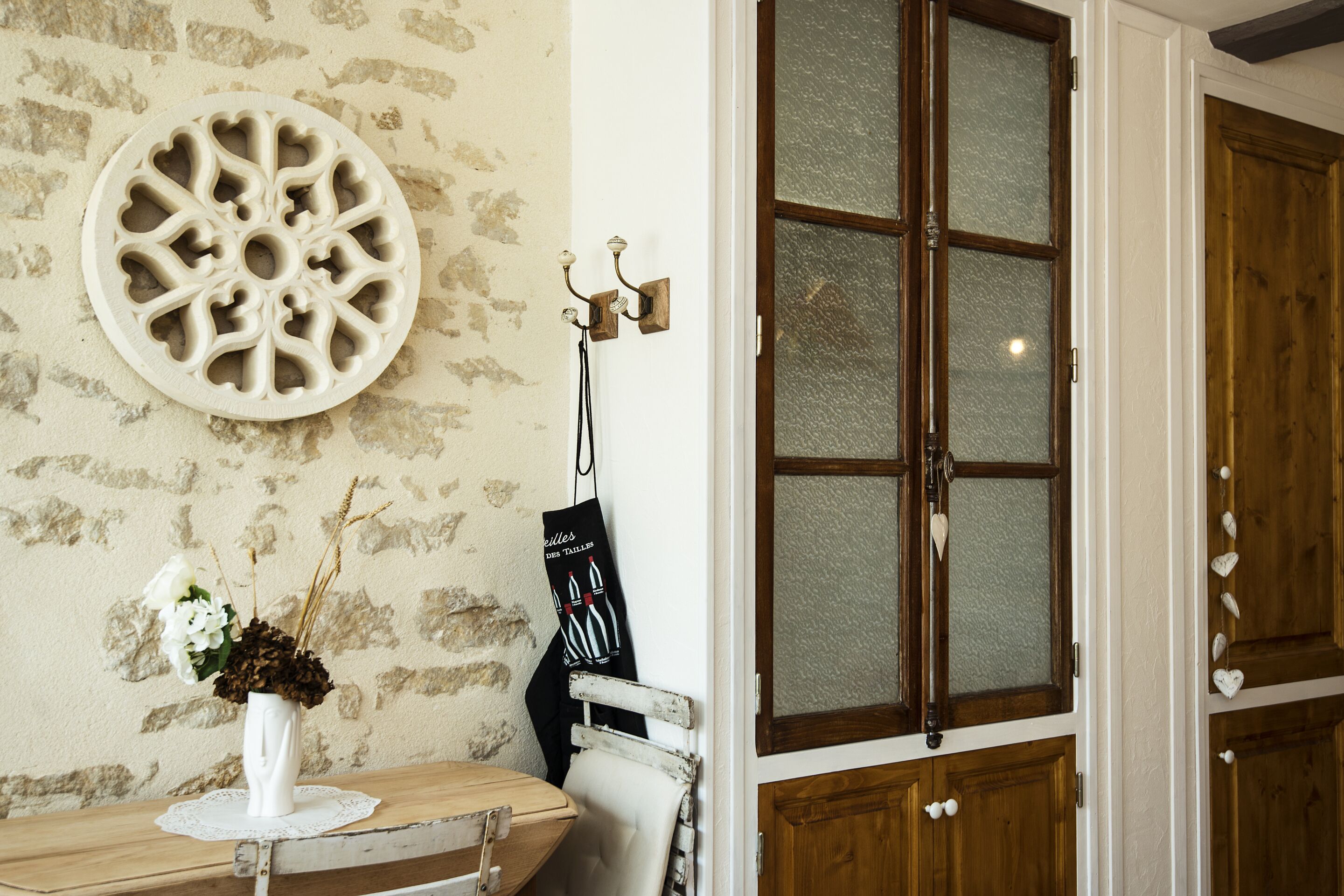 Una stanza con una parete in pietra rustica, un piatto decorativo rotondo, un vaso bianco con fiori, una sedia di legno e una porta di legno con pannelli in vetro.