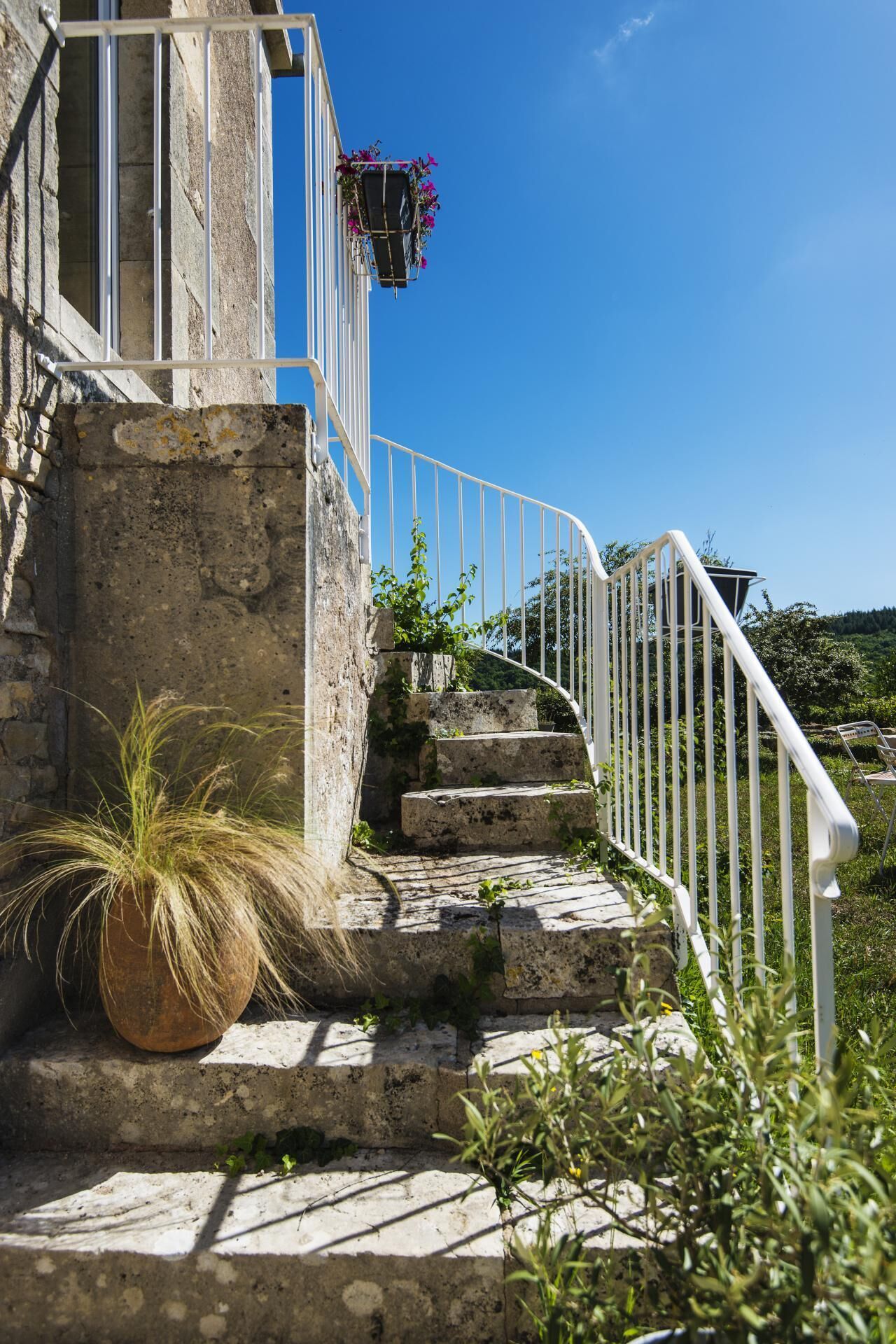 A stone staircase leading to a building with a white railing, surrounded by greenery and a clear blue sky.