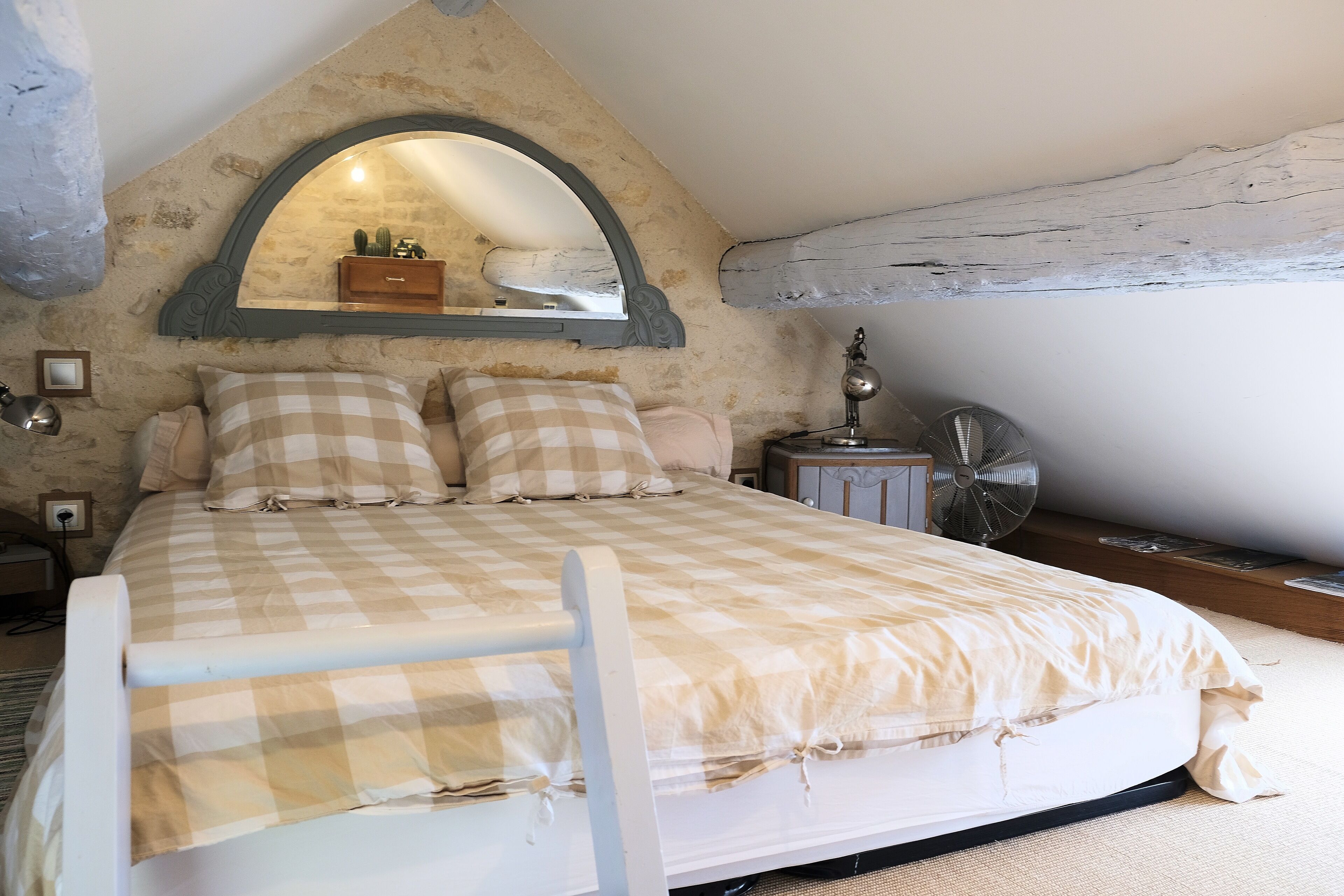 Un dormitorio con una colcha a cuadros, un espejo sobre la cama, una lámpara de noche y un ventilador en el suelo.