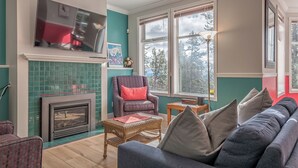 A living room with a fireplace, a television mounted above it, a large window with a view of trees, and a sofa with cushions. - Large suite on the Knoll in a cul-de-sac w/ a private hot tub and pet friendly! (Silver Star Mountain)