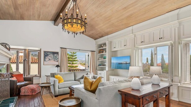 A spacious living room with a wooden ceiling, a chandelier, a sofa set, a TV, and a view of the sea through large windows. - Superb La Vida Riviera 2 BD 2BA Gorgeous Riviera Views, Walk Everywhere! (Santa Barbara)