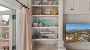 A room with a built-in shelf unit displaying decorative items, a framed picture, and a view of a coastal town. - Superb La Vida Riviera 2 BD 2BA Gorgeous Riviera Views, Walk Everywhere! (Santa Barbara)