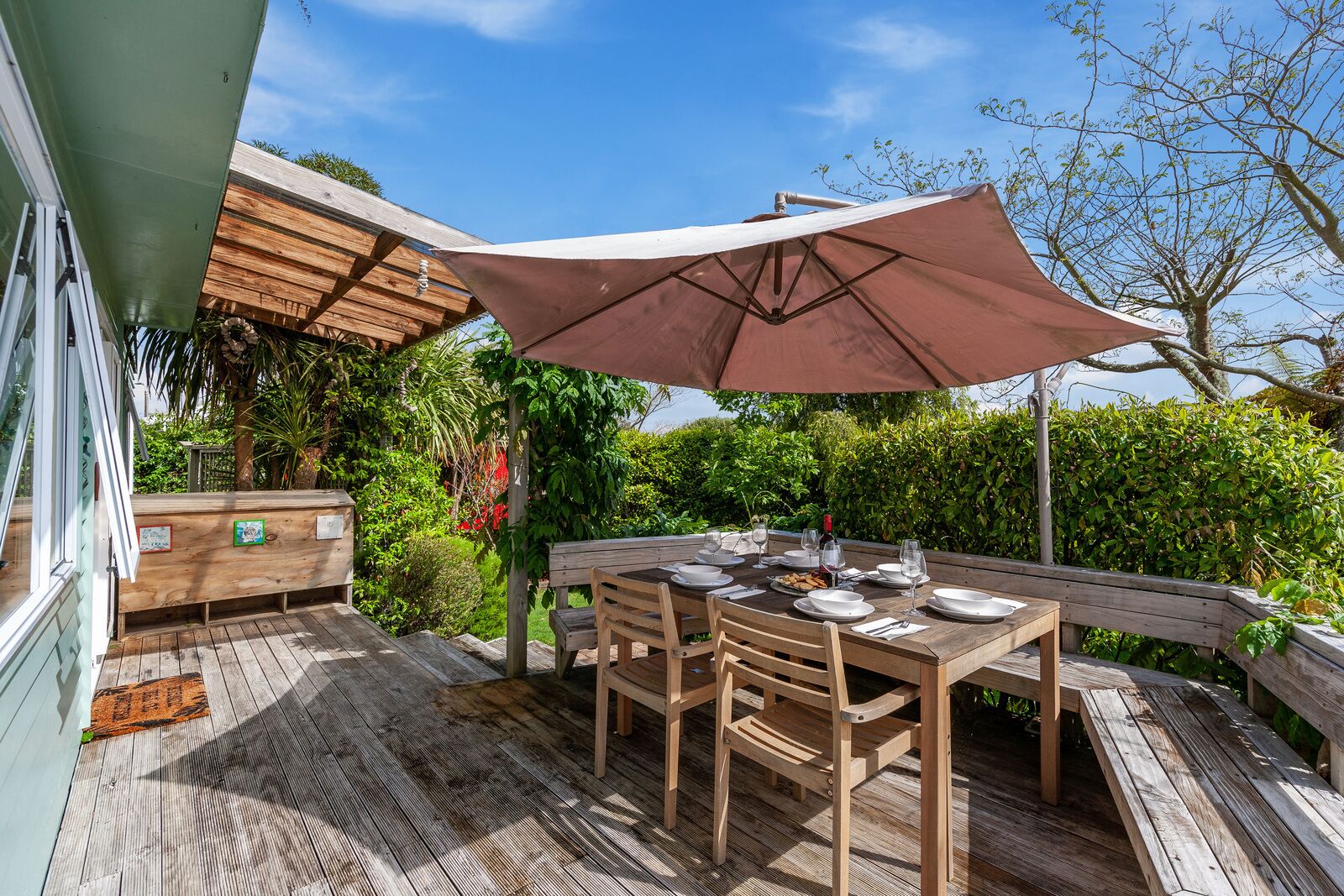 Een terras van hout buiten, met een eettafel, omringd door weelderige begroeiing en een heldere lucht.