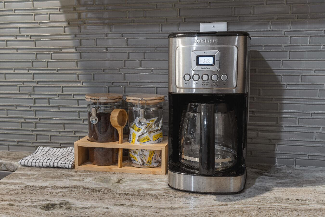 Une machine à café dotée d’un écran numérique, une cuillère en bois et un bocal en verre contenant des sachets de thé, le tout posé sur un plan de travail de cuisine.