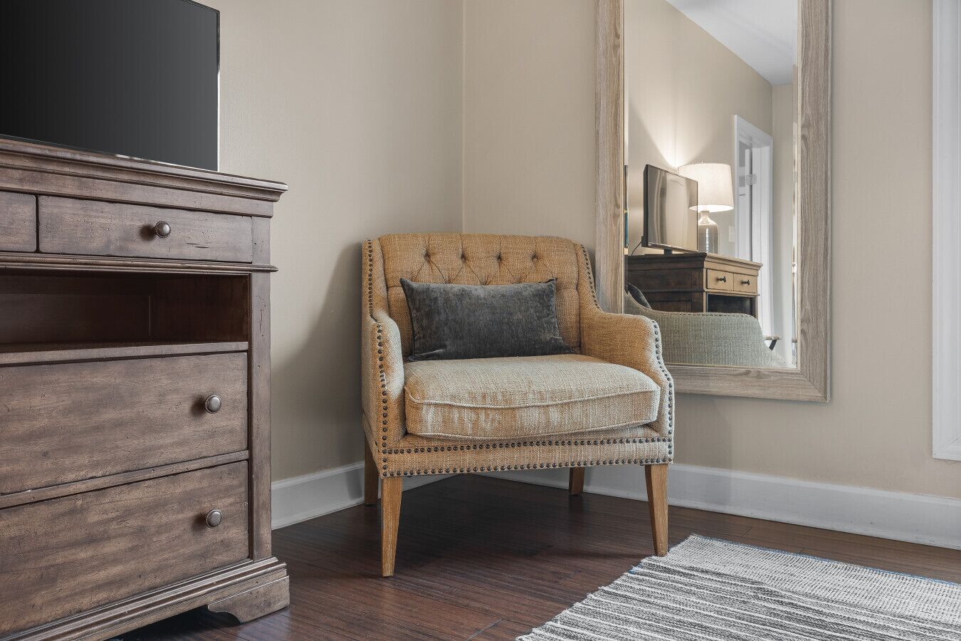 A corner room with a wooden dresser, a cushioned armchair, and a large mirror on the wall.