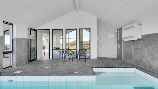 Ein moderner Poolbereich im Innenbereich mit freiem Blick auf den Pool, einer Lounge mit Stühlen und einem großen Fenster, das natürliches Licht hereinlässt.