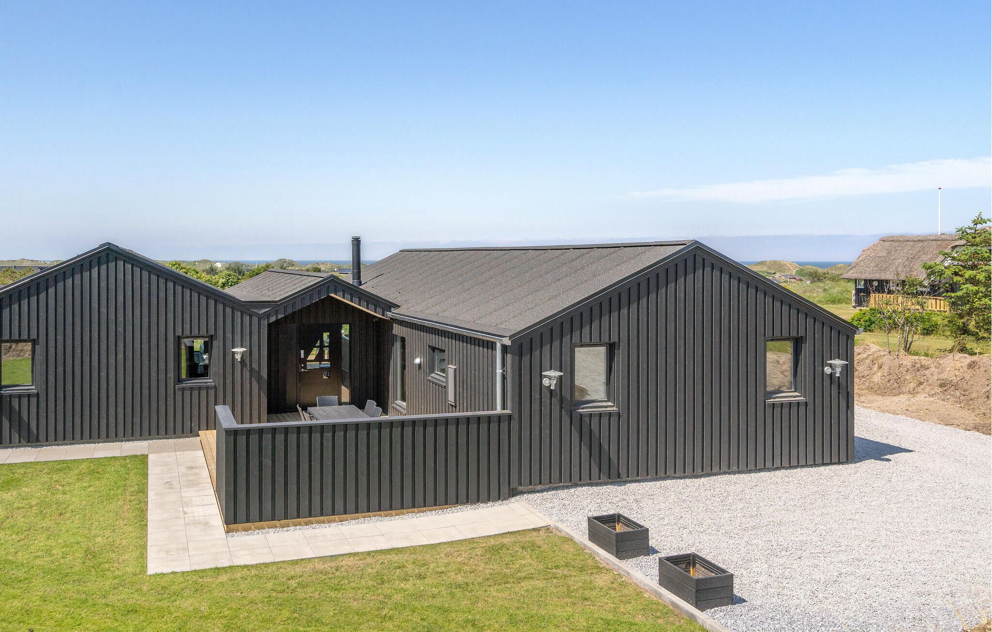 Een modern gebouw in donkere tinten, met een schuin dak, een terras met zitplaatsen en een grindpad.