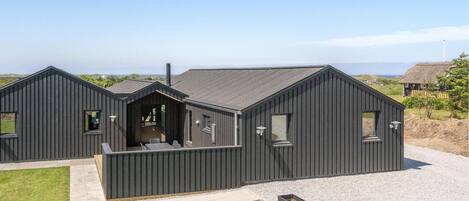 Een modern gebouw in donkere tinten, met een schuin dak, een terras met zitplaatsen en een grindpad.
