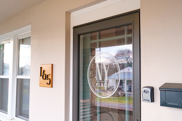 A building entrance with a glass door displaying a circular logo with "NY PLACE" and "Harrisonburg, Virginia" text. A wooden plaque with the number "165" is mounted on the wall.