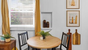 A dining area with a round wooden table, two black chairs, a wooden sideboard, and a window with white blinds and yellow curtains. - SD Getaway - Boho Studio, Spacious Yard w/ BBQ (San Diego)