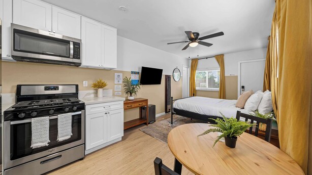 A compact kitchen with a microwave, stove, and cabinets. There's a dining table with a plant, a bed with a ceiling fan, and a TV on the wall. - SD Getaway - Boho Studio, Spacious Yard w/ BBQ (San Diego)