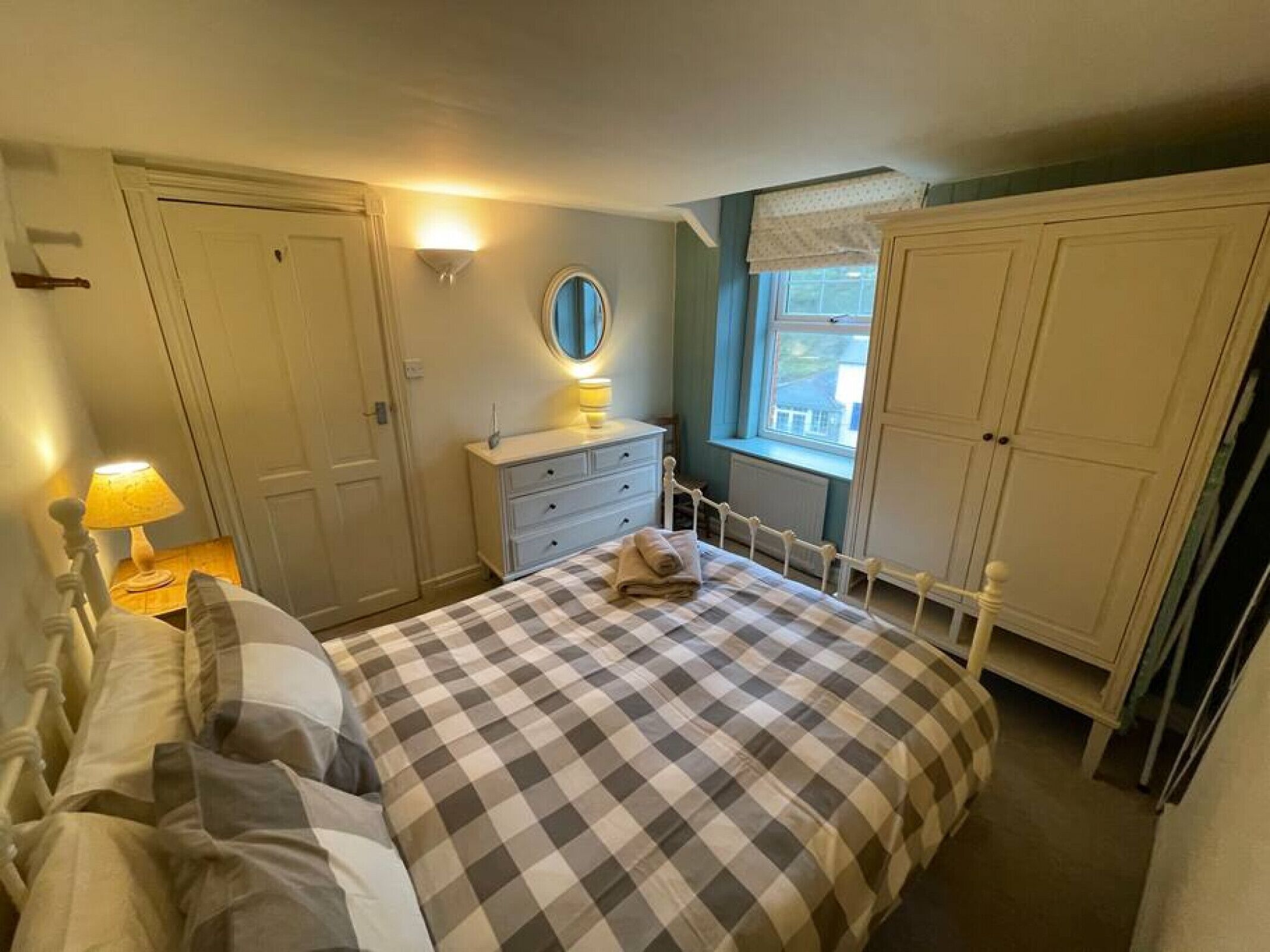 Un dormitorio con una colcha a cuadros, una lámpara de noche, un aparador, un espejo y una ventana con cortinas.
