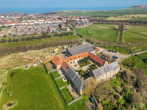 An aerial view of a large estate with multiple buildings, including a main house with a red-tiled roof, surrounded by well-maintained gardens and fields.