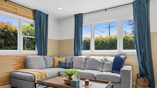 A modern living room with a grey sectional sofa, a wooden accent wall, a coffee table, and a view of greenery through large windows.