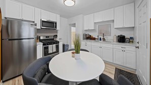 A modern kitchen with white cabinets, a stainless steel refrigerator, a microwave, and a stove. There is a round table with two chairs and a potted plant.