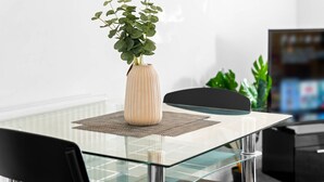 Ett modernt matområde med ett glasbord, en beige vas med gröna växter och en svart stol.