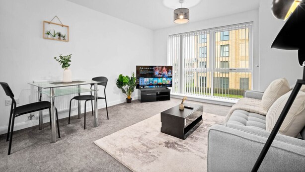 Ett modernt vardagsrum med ett matbord i glas, svarta stolar, en grå soffa, ett soffbord och en tv. Det finns två hänglampor, en inramad tavla på väggen och en växt på bordet.