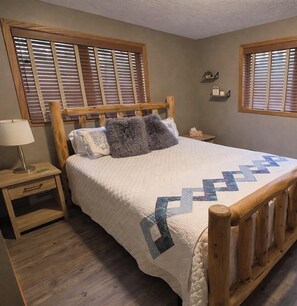 A bedroom with a wooden bed, a bedside table with a lamp, and a window with blinds.