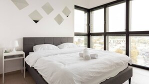 Moderni hotellihuone, jossa on suuri sänky, yöpöytä ja näkymä suurista ikkunoista.