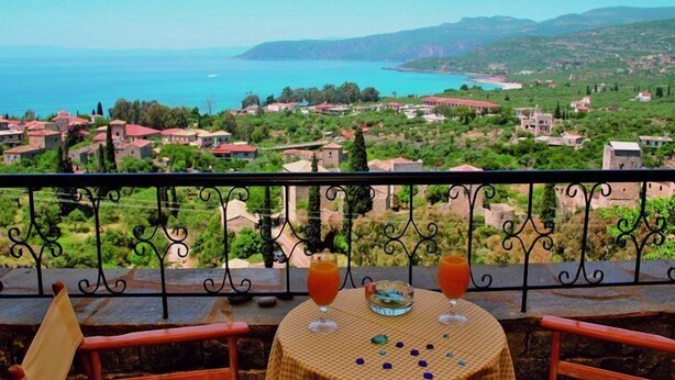 A balcony with a table set for two, overlooking a coastal town with houses and a view of the sea.