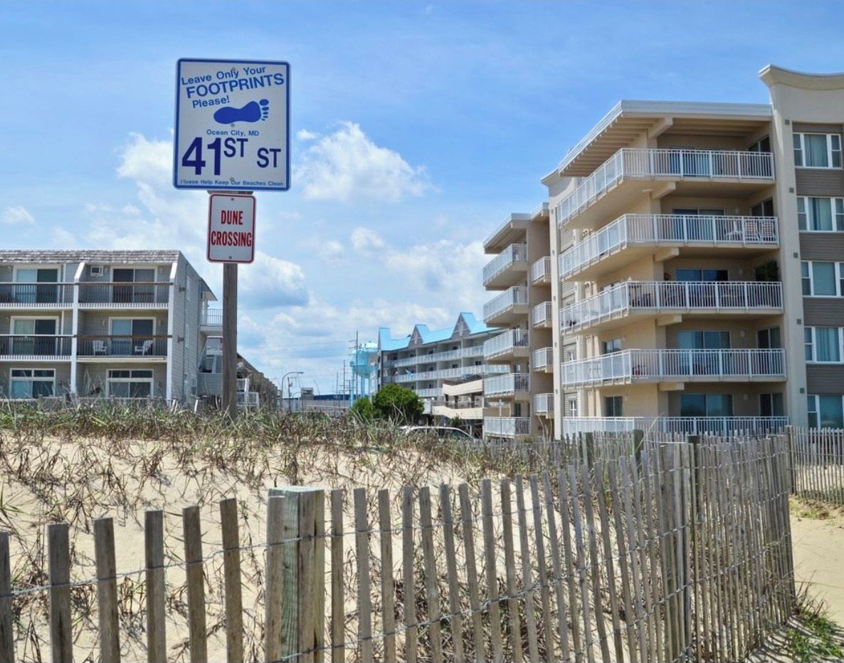 Un bâtiment situé en bord de mer, doté de balcons, une pancarte indiquant : « Ne laissez que vos empreintes, s’il vous plaît ! », et un panneau indiquant : « Passage des dunes ».