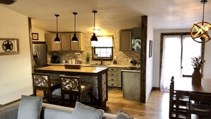 A modern kitchen with wooden cabinets, a central island, pendant lights, and a dining area with a wooden table and chairs. - Nolia Cabin in the Little River Valley (Nashoba)