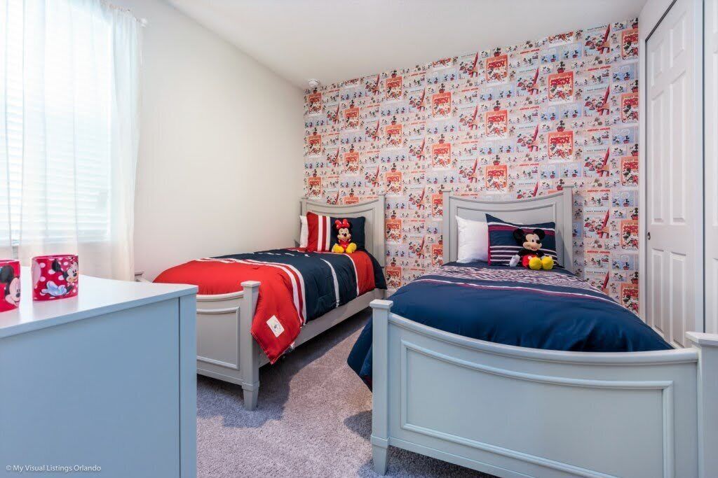 Une chambre avec deux lits, un bureau et une fenêtre. Les murs sont recouverts d’un papier peint sur le thème de Mickey Mouse.