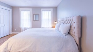 Un dormitorio con una cama grande, dos ventanas con cortinas translúcidas, una lámpara de noche y una puerta.